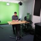 Man in plaid shirt at desk in small green-screen studio with lights and camera